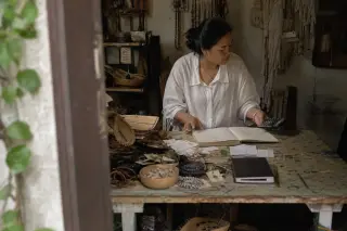 Craftswoman in a small city workshop reviewing her ledger with handmade goods on her desk, natural light and candid atmosphere.