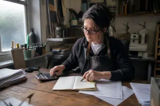 Small business owner in a craft workshop reviewing a paper ledger and invoices in natural morning light, showing focus and authentic workspace details.