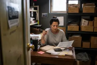 Small business owner in a cluttered storage room, reviewing financial documents with natural morning light from a high window.