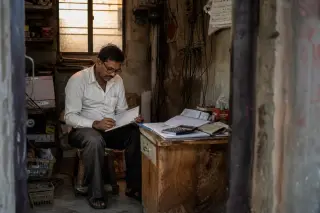 Artisan electrician seated in a workshop, reviewing handwritten financial notes at dusk, with gentle sunlight illuminating a workbench and scattered tools.