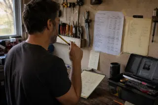 Electrician in a modest garage workshop, focused on analyzing handwritten notes and a printed financial sheet, with natural light filtering through a side window.