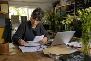 Florist in a modest workspace reviewing finances with handwritten notes, invoices, and a laptop, natural light adding warmth and realism.