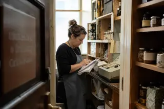Shop owner reviewing receipts and notes in a storeroom, illuminated by soft daylight, with shelves of goods in the background.